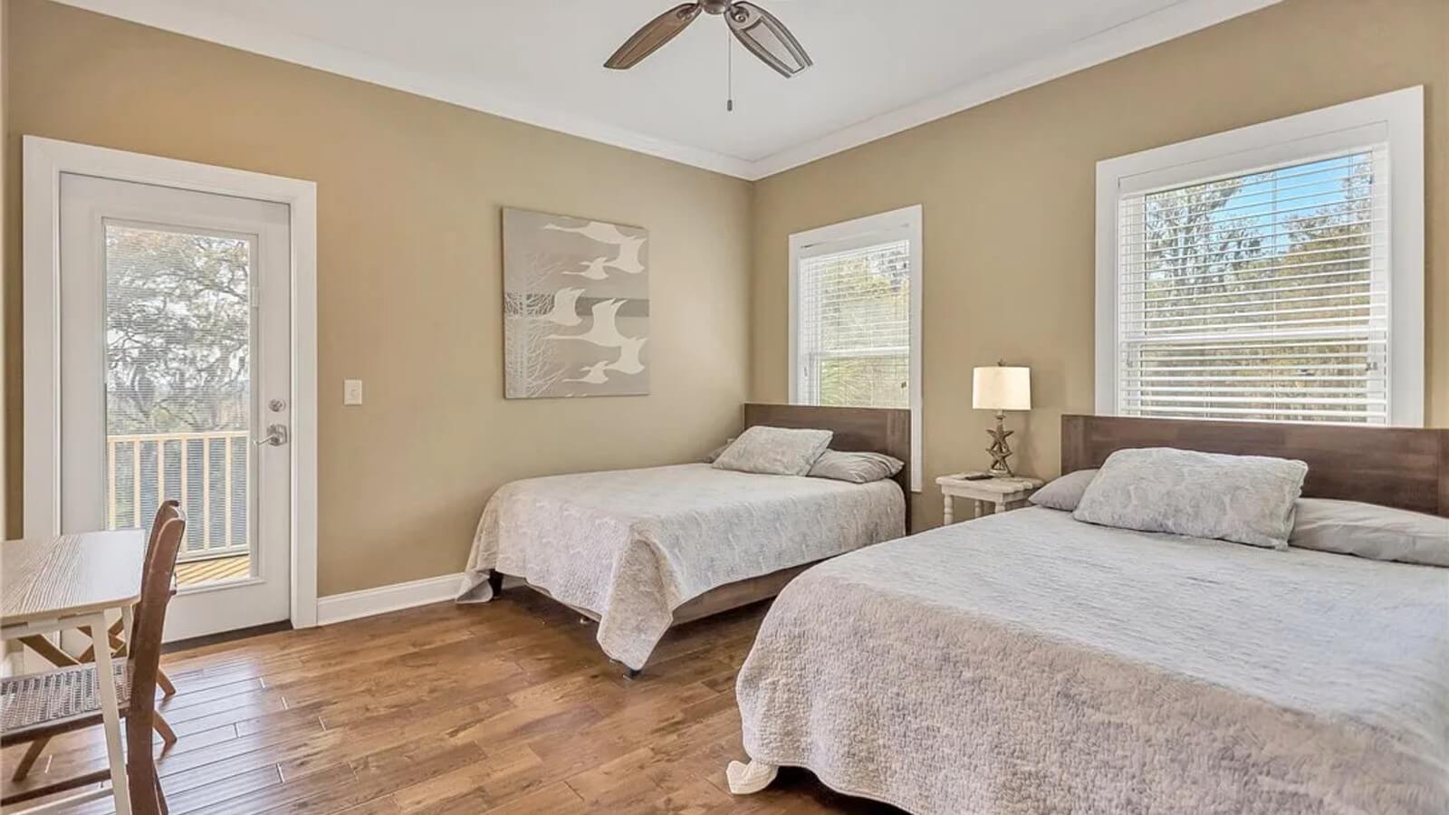 Bedroom with two wooden beds and neutral bedding, a nightstand with a lamp between them, and a balcony door on the left. A ceiling fan and beige walls complete the room.