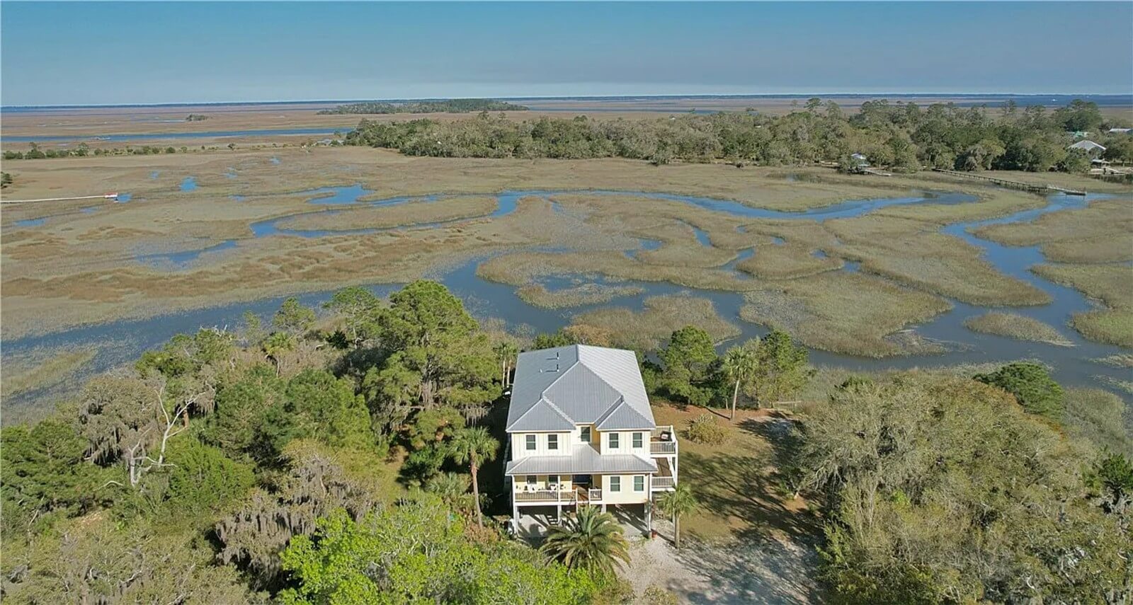 A light-colored house on stilts nestled among trees, overlooking a expansive marsh with winding inlets and shallow water channels.