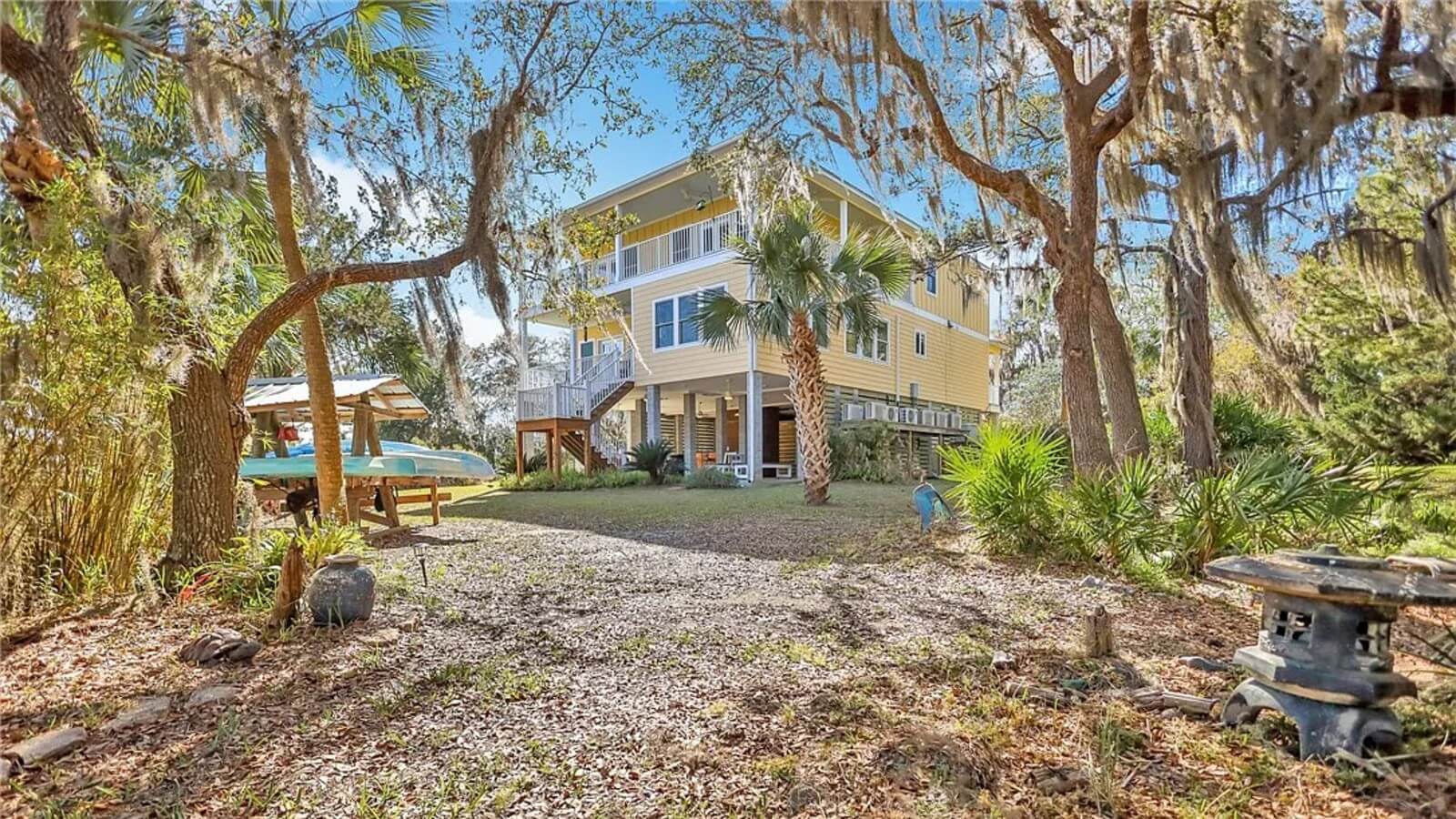 Yellow two‑story house on stilts in a sunlit wooded yard with palm trees and a gravel clearing under the home's deck.