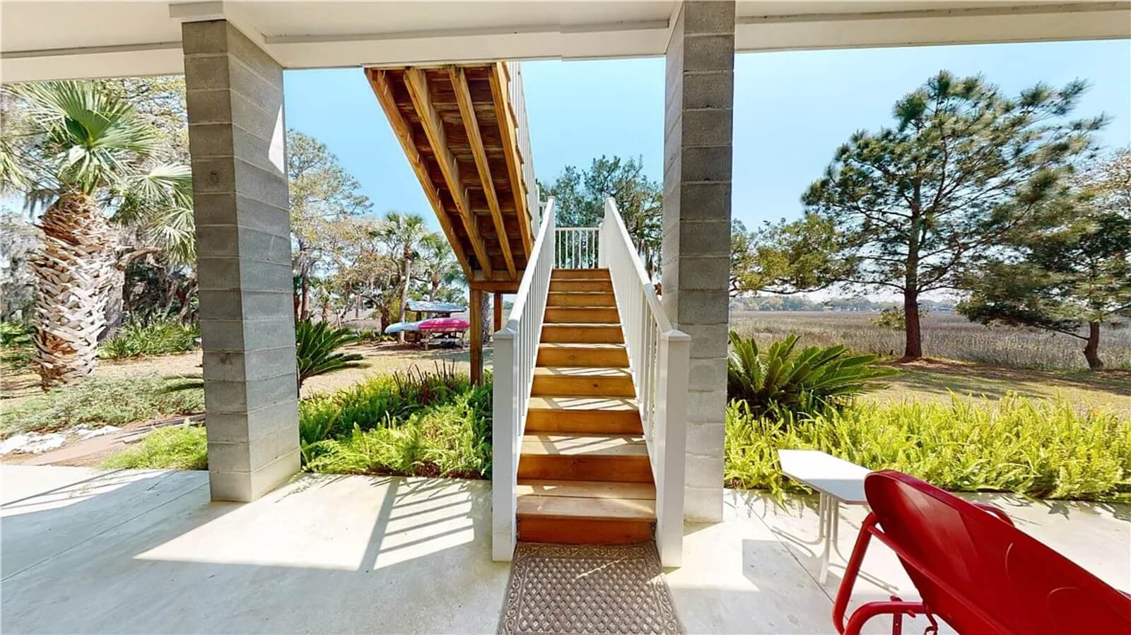 Outdoor patio with a wooden staircase up to a raised deck, surrounded by palm trees and greenery under a sunny sky.