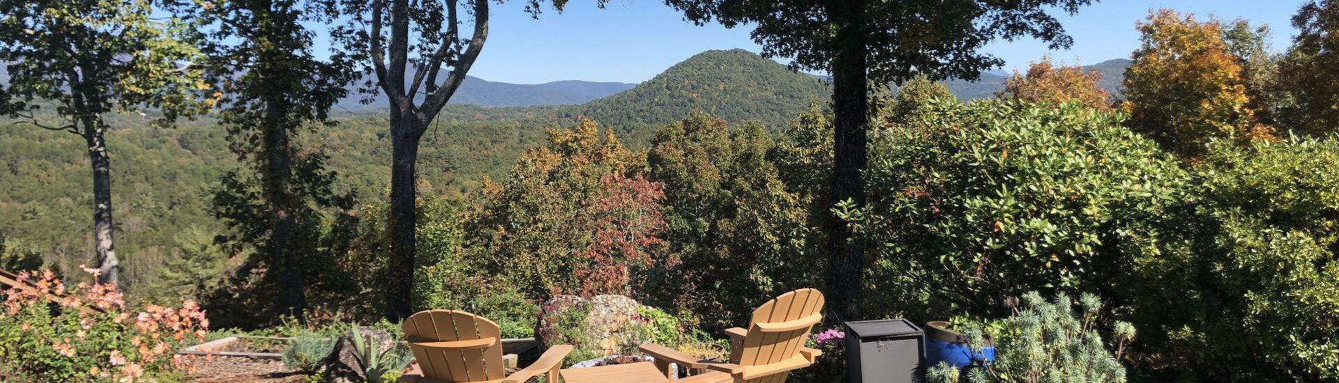 Two light tan Adirondack chairs on brick patio with fire pit surrounded by flower garden, bushes, and trees looking out to tree-covered hills