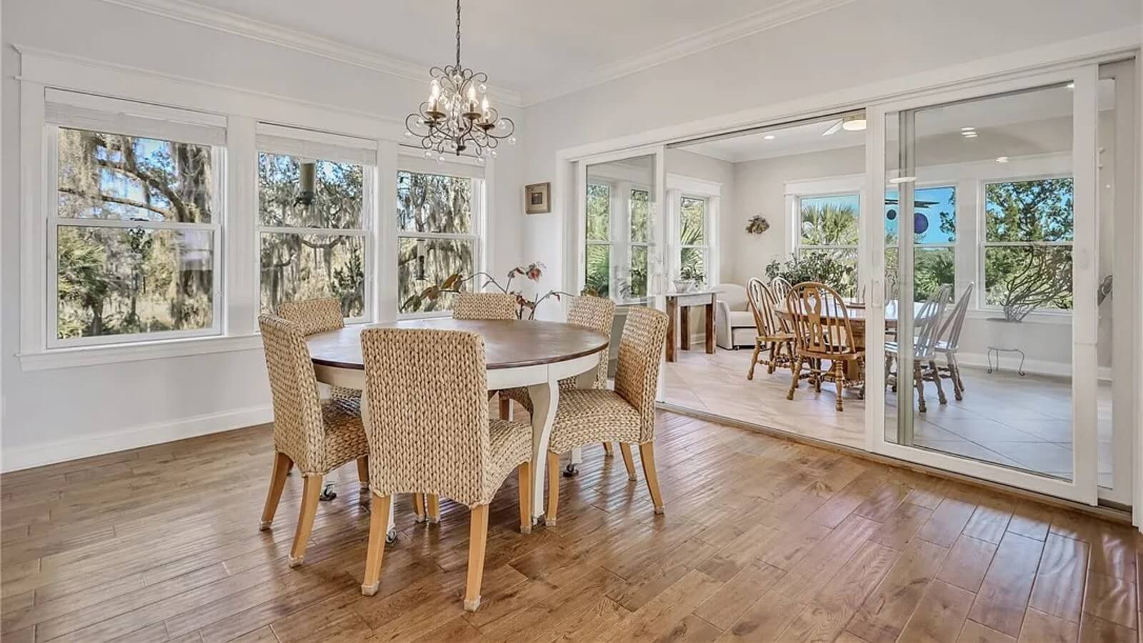 Bright dining area with a round wooden table and wicker chairs on casters, a crystal chandelier, and large windows; sliding glass doors to another room artfully separate spaces.
