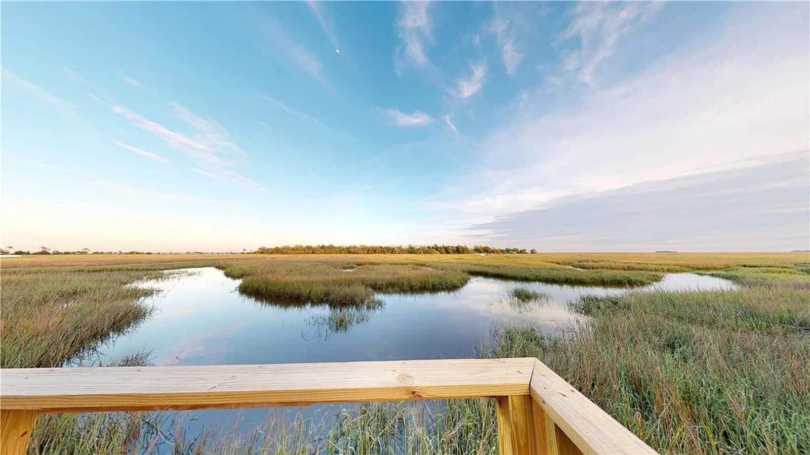 Wooden railing of a boardwalk over a calm marsh with grasses and reflective water under a blue sky.