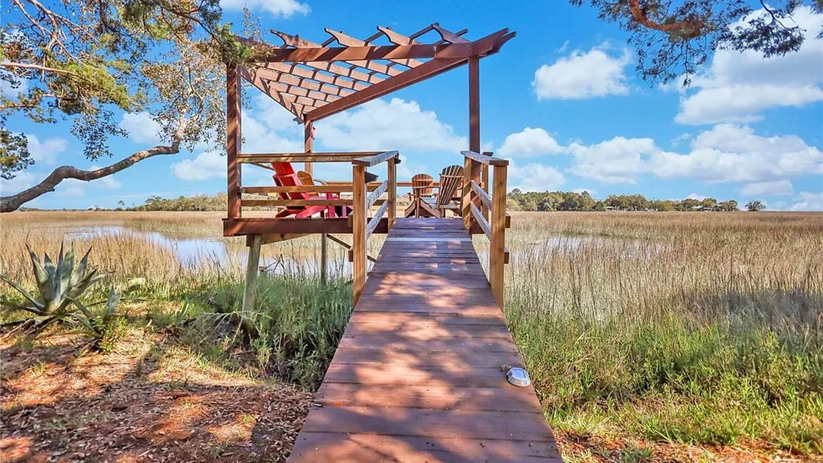 Wooden boardwalk leading to a small viewing deck with red Adirondack chairs under a pergola, over a marshy field on a sunny day.