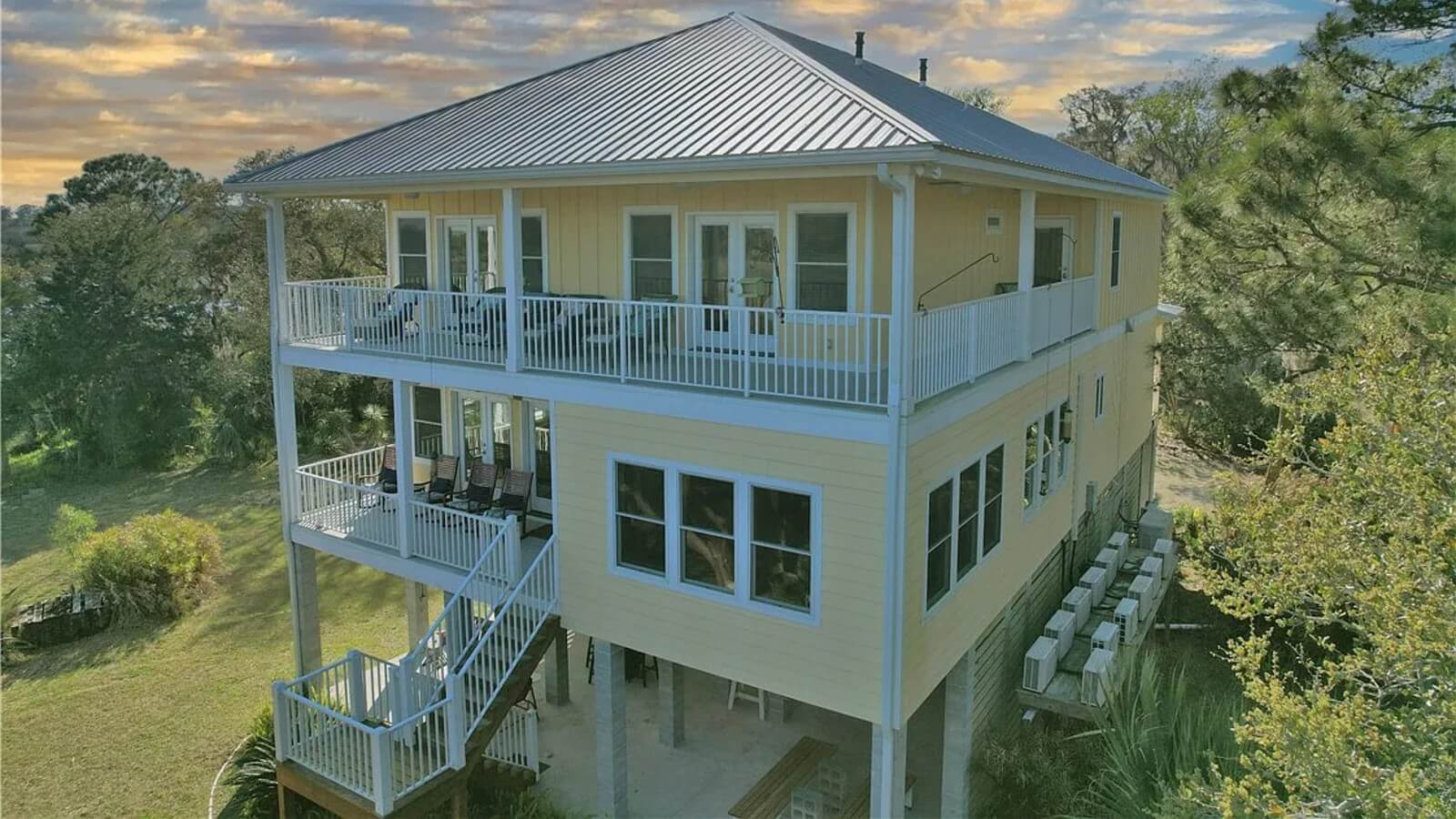 Three-story yellow house on stilts with white railings and wraparound decks, metal roof, surrounded by trees at sunset.