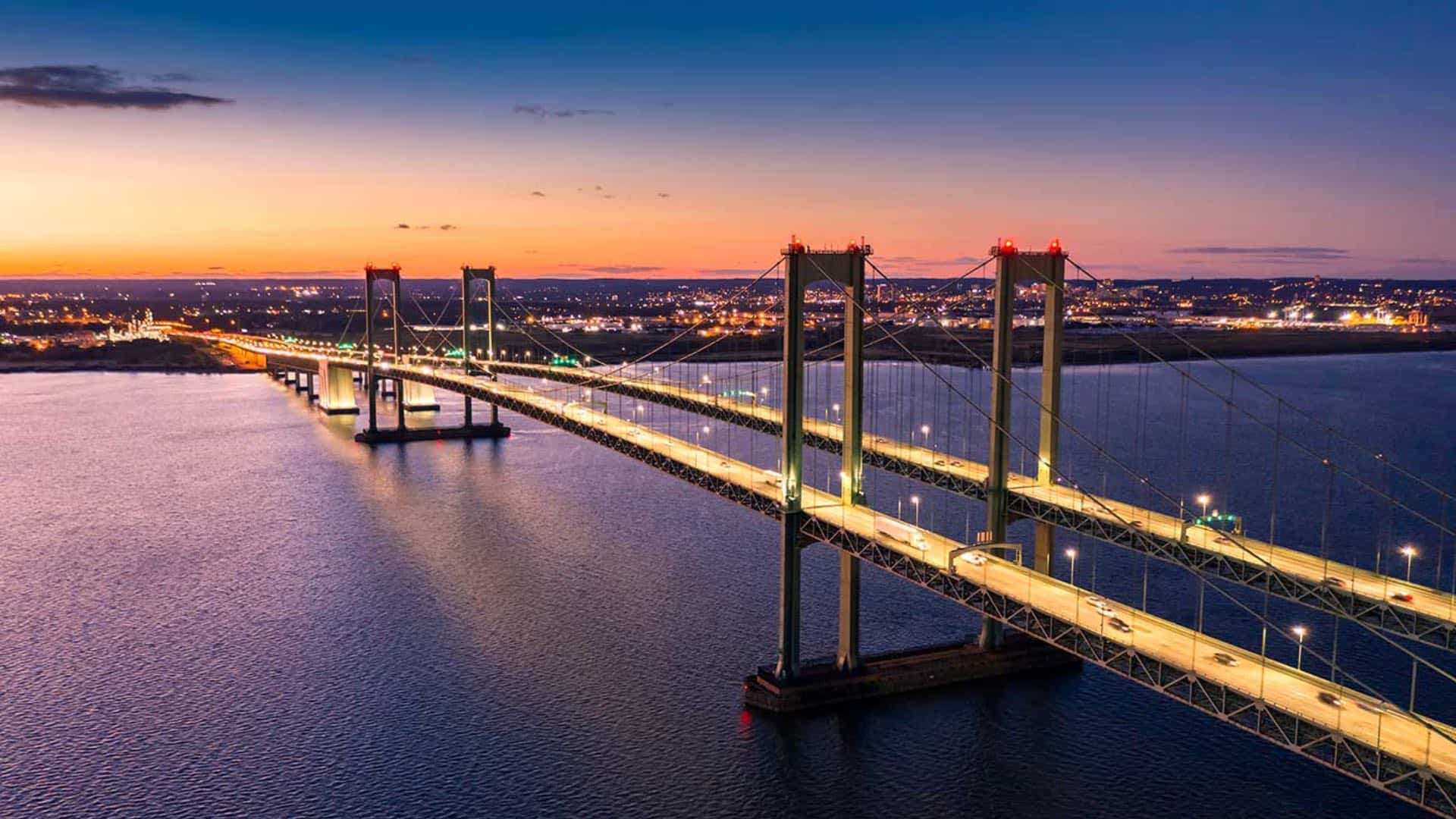 Large lighted up bridge across a large body of water with a city in the background at dusk